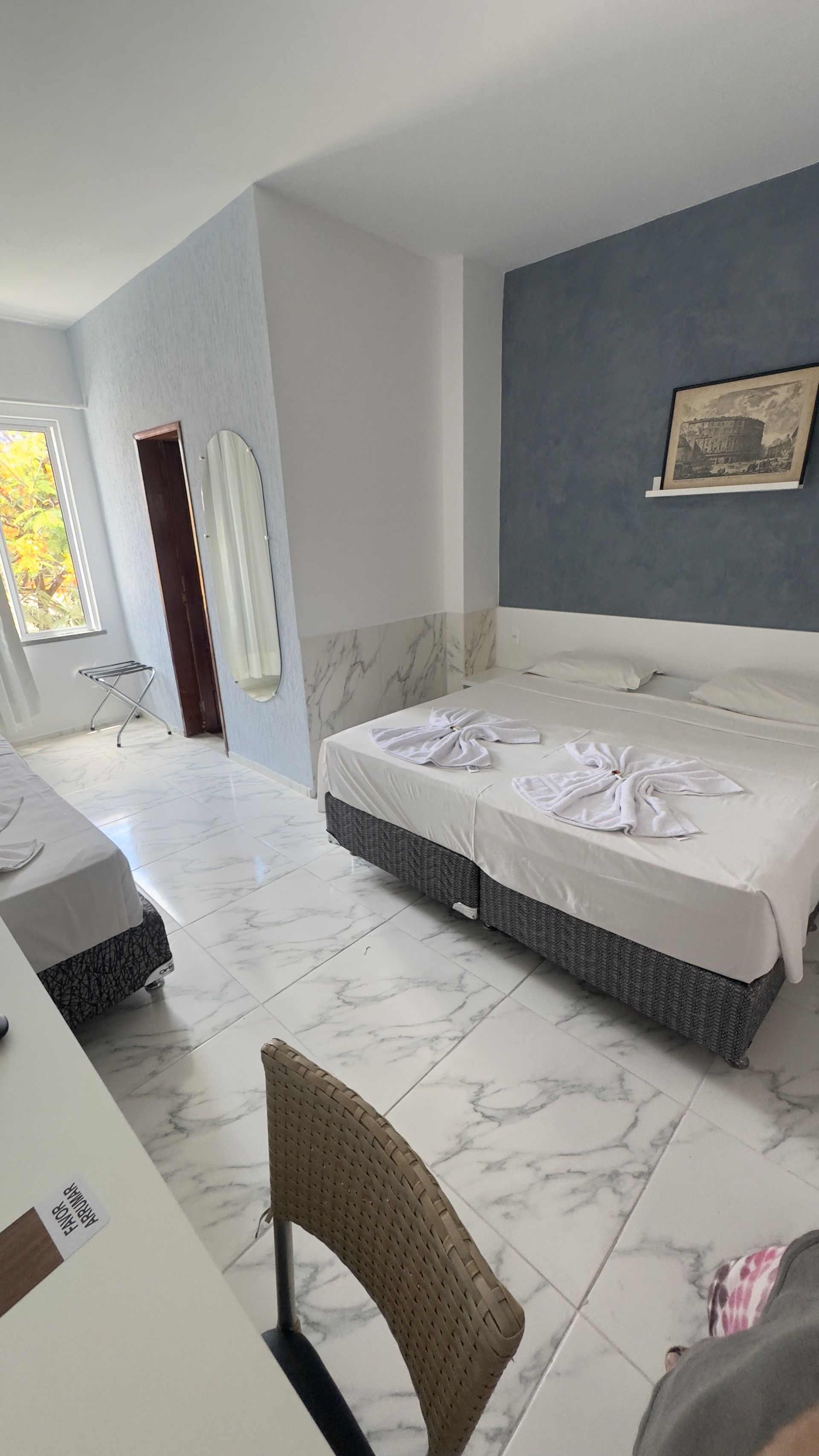 Quarto de hotel luminoso com cama, piso de mármore, parede com detalhes em azul e uma pequena mesa junto à janela.
