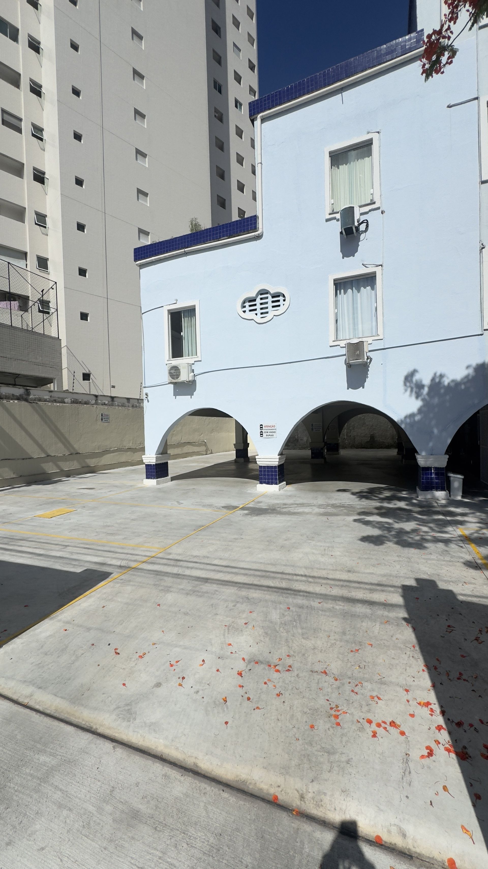 Prédio branco com aberturas em arco e detalhes em azul ao lado de um pátio pavimentado, próximo a um prédio alto de apartamentos.