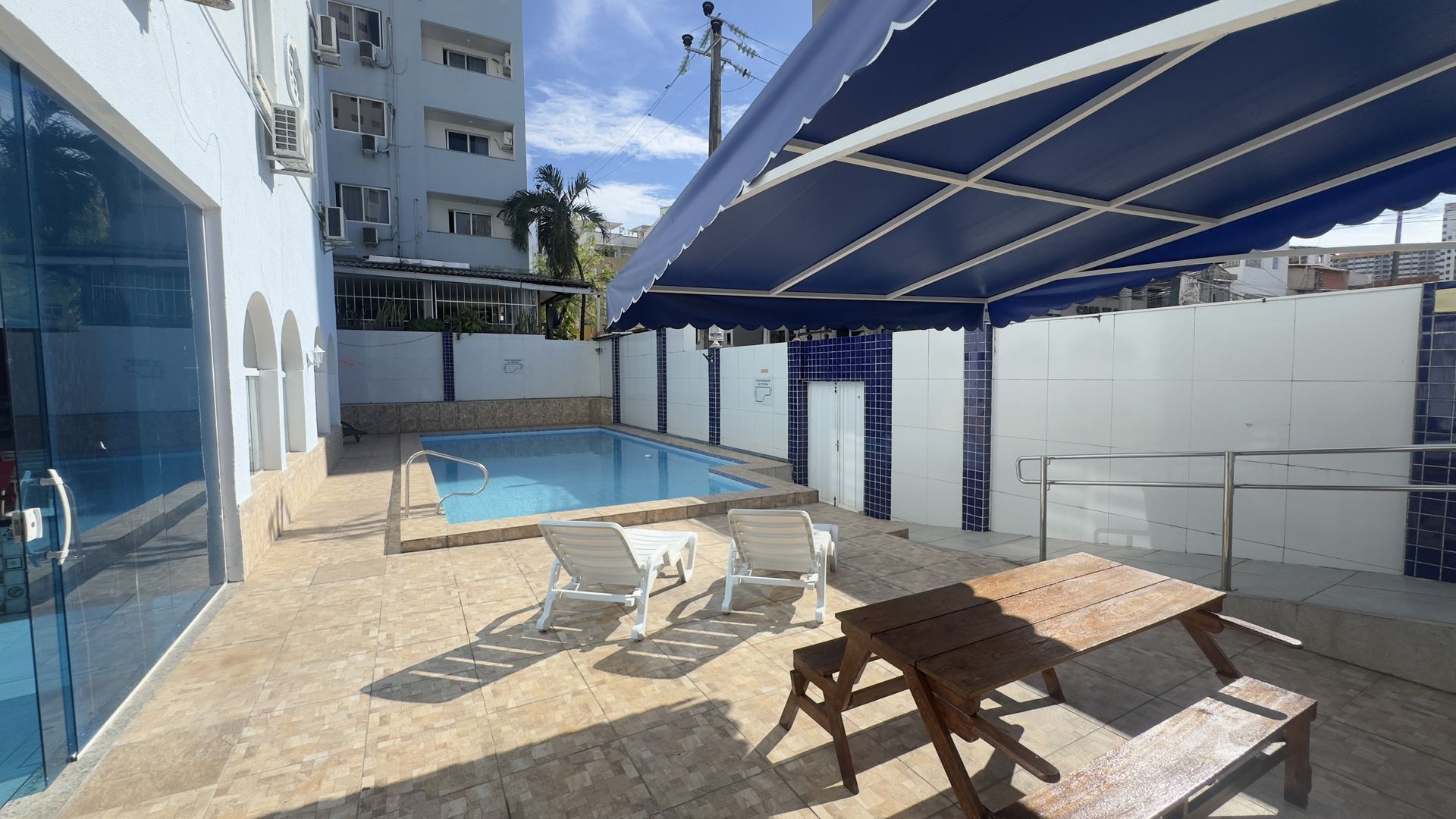 Pátio à beira da piscina com espreguiçadeiras, mesa de piquenique e toldo azul ao lado de um edifício.