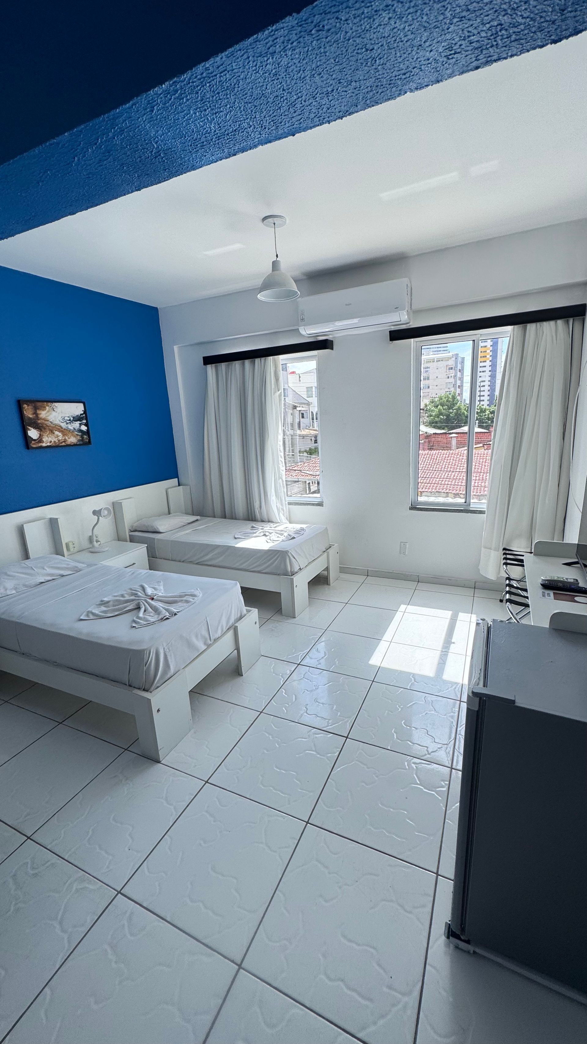 Quarto moderno e luminoso com duas camas, parede de destaque em azul, piso de azulejo branco e janelas amplas com cortinas.