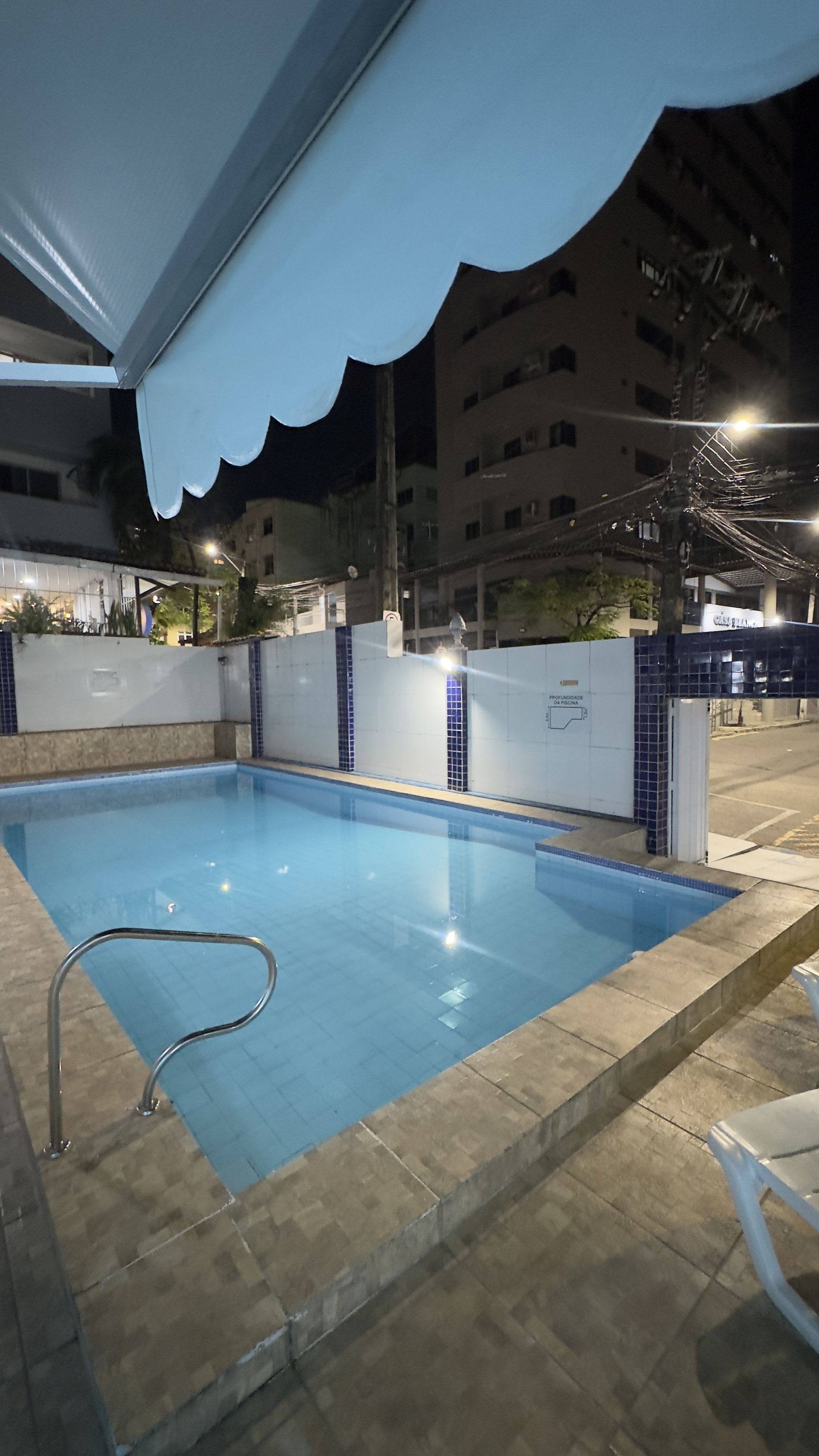 Pequena piscina na cobertura à noite, com água azul, paredes brancas e um prédio de apartamentos iluminado ao fundo.
