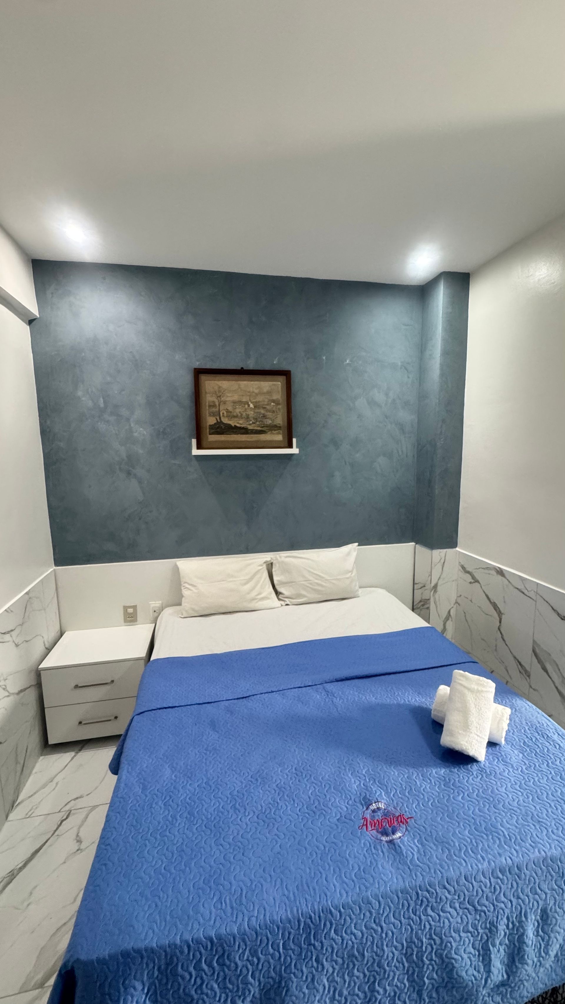 Quarto pequeno com parede de destaque em azul, cama com cobertor azul, travesseiros brancos e obra de arte emoldurada acima da cabeceira.