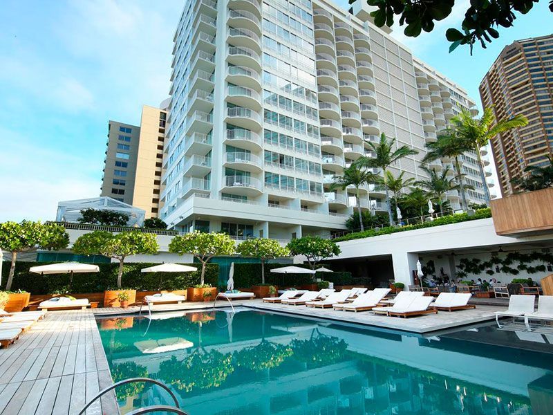 Piscina de resort com espreguiçadeiras brancas ao lado de um hotel alto sob um céu azul.