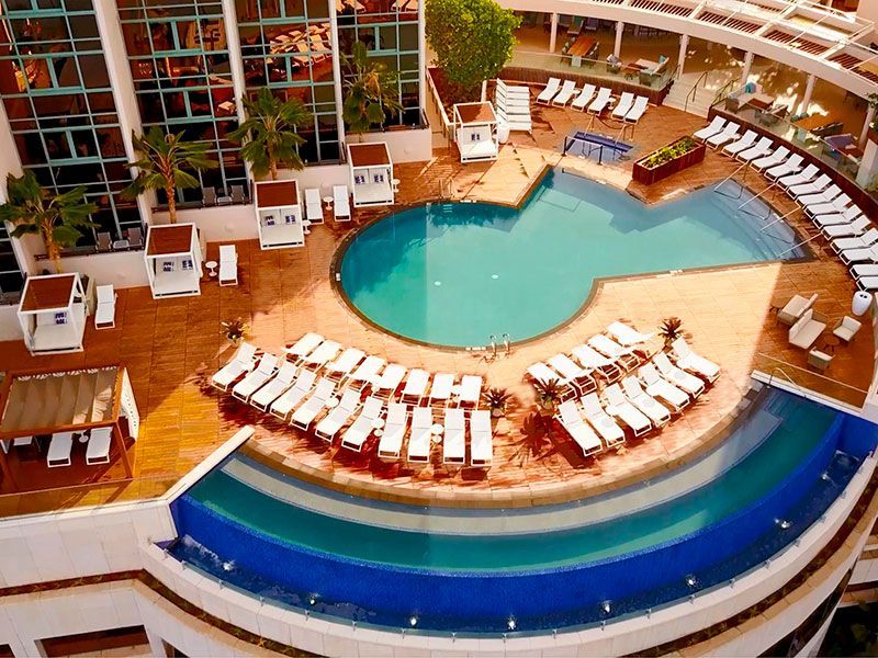 Vista aérea da piscina de um resort com água azul, espreguiçadeiras brancas e um deck curvo sob um toldo colorido.