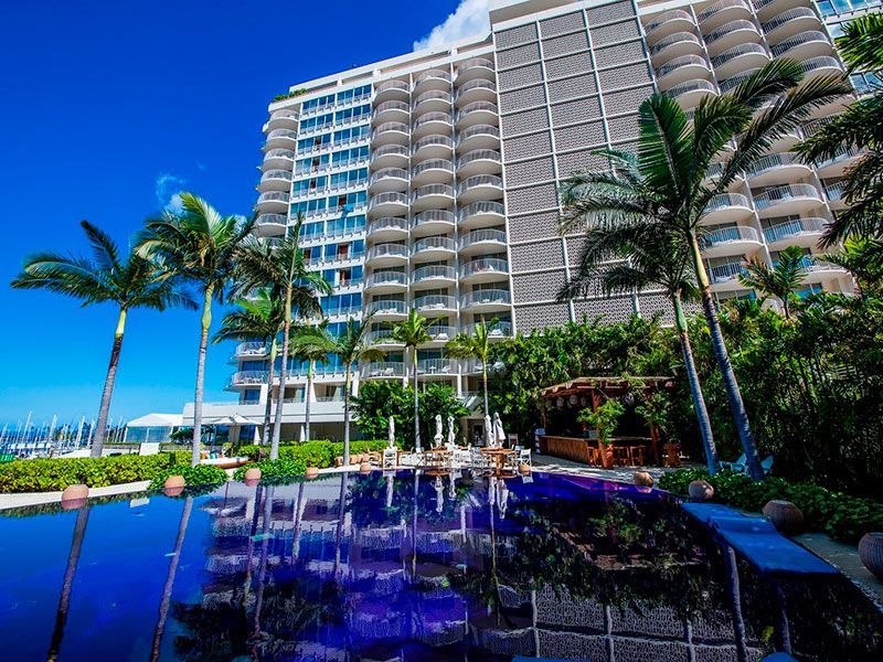 Piscina de resort tropical com palmeiras e um alto hotel à beira-mar sob um céu azul brilhante.