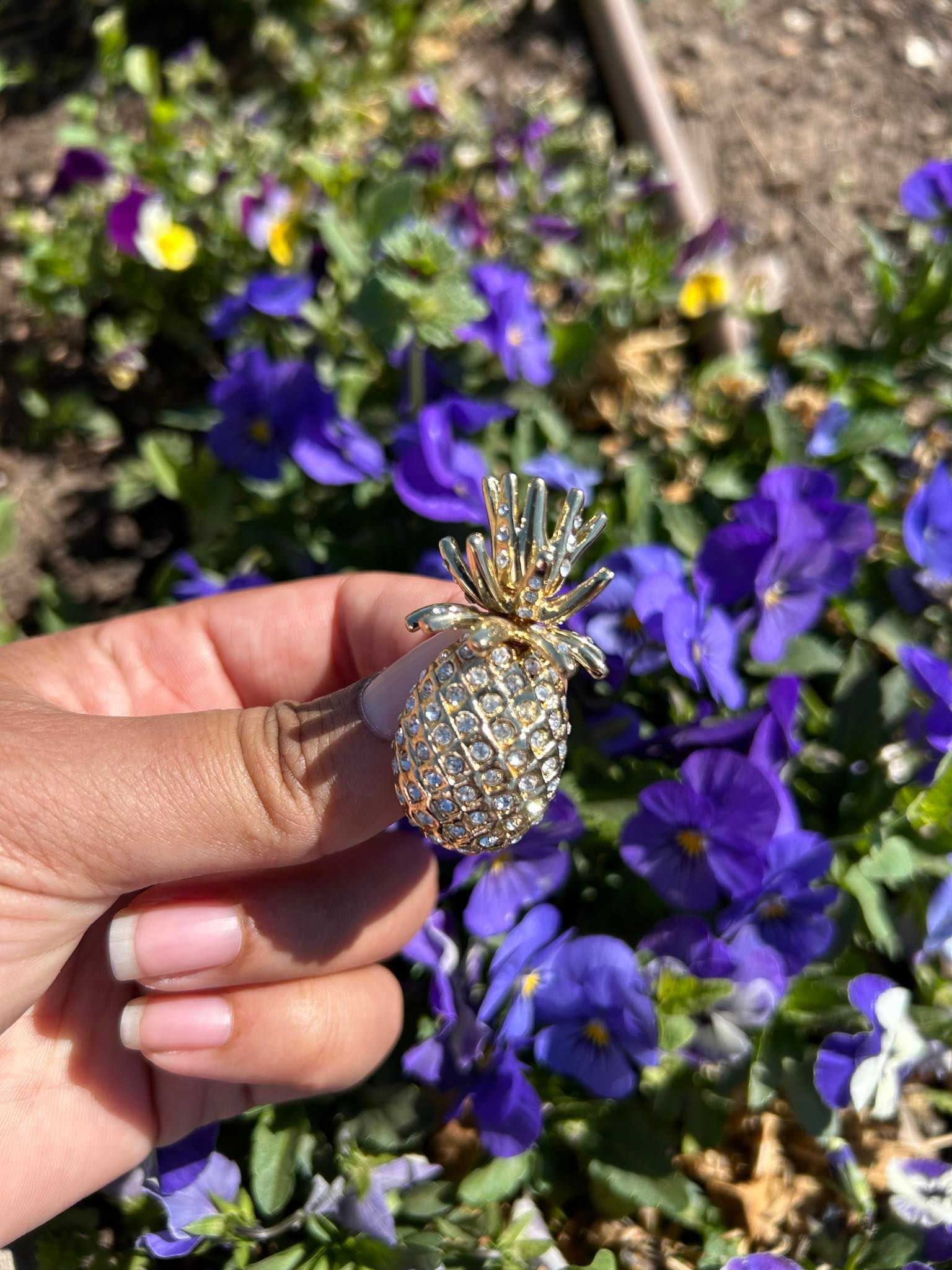 A person is holding a pineapple brooch in front of purple flowers.
