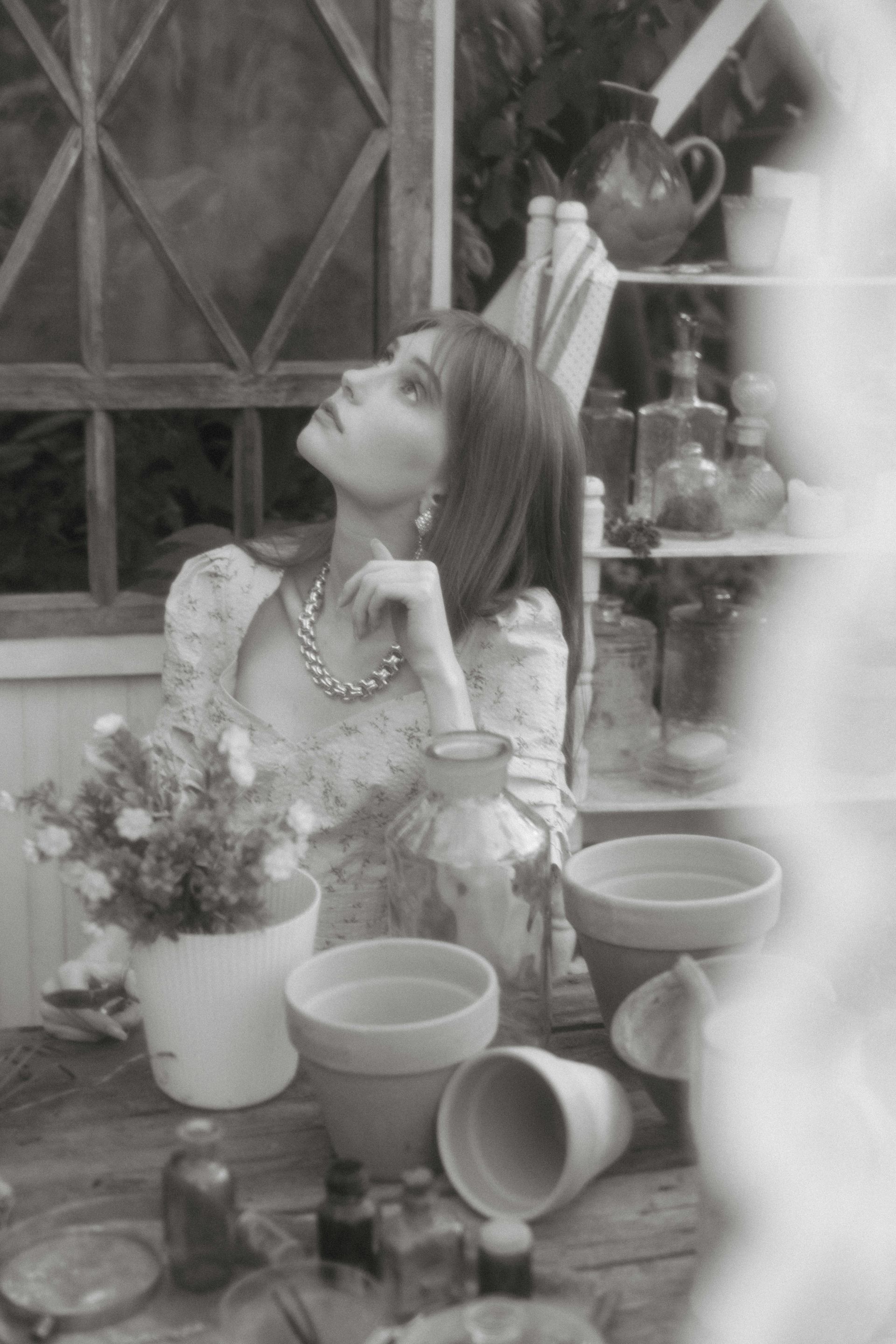 A black and white photo of a woman sitting at a table with potted plants.