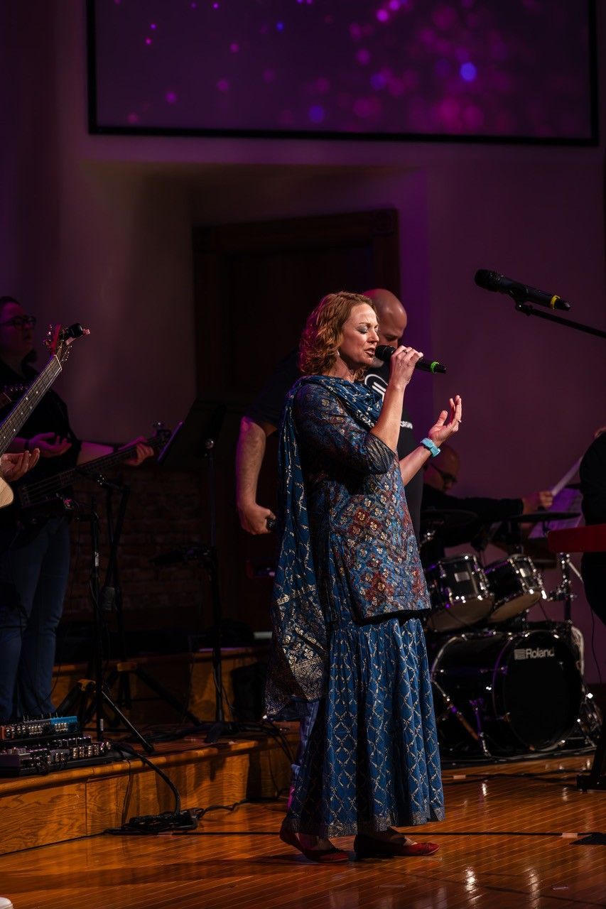 A singer in a patterned blue outfit performs with a microphone on a stage with musical instruments in the background.