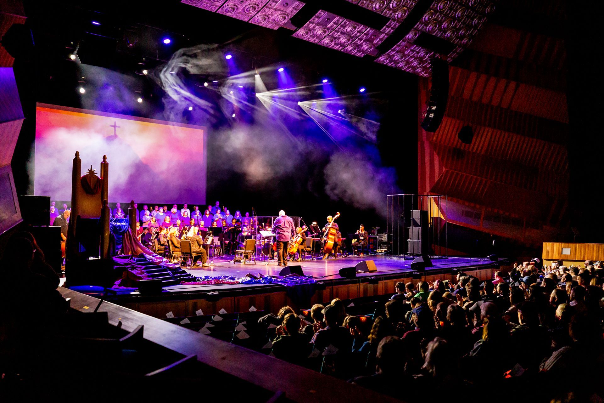 A conductor leads an orchestra on a brightly lit stage in a theater, with a large screen in the background and an audience.