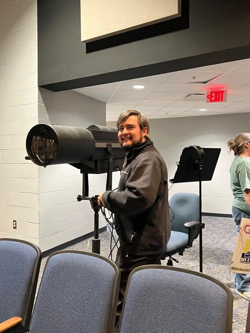 A person standing in a theater auditorium behind a stage spotlight, smiling at the camera.