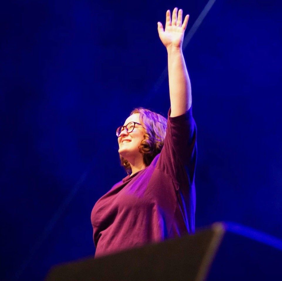 A person with glasses and purple-toned hair in a purple shirt, smiling and holding one arm up against a dark blue background.