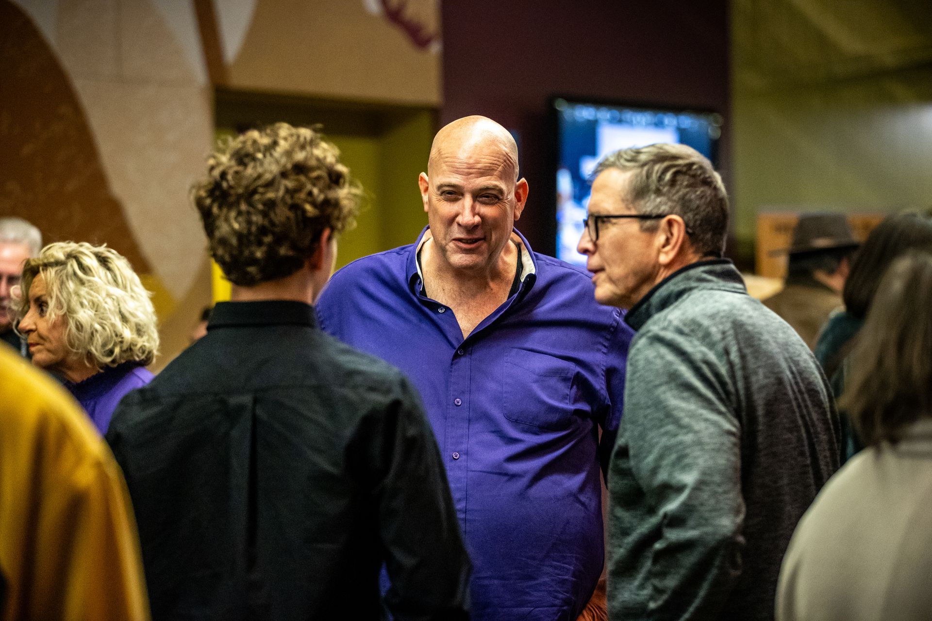A group of people standing in an indoor space, with a man in a blue button-down shirt speaking to two others.