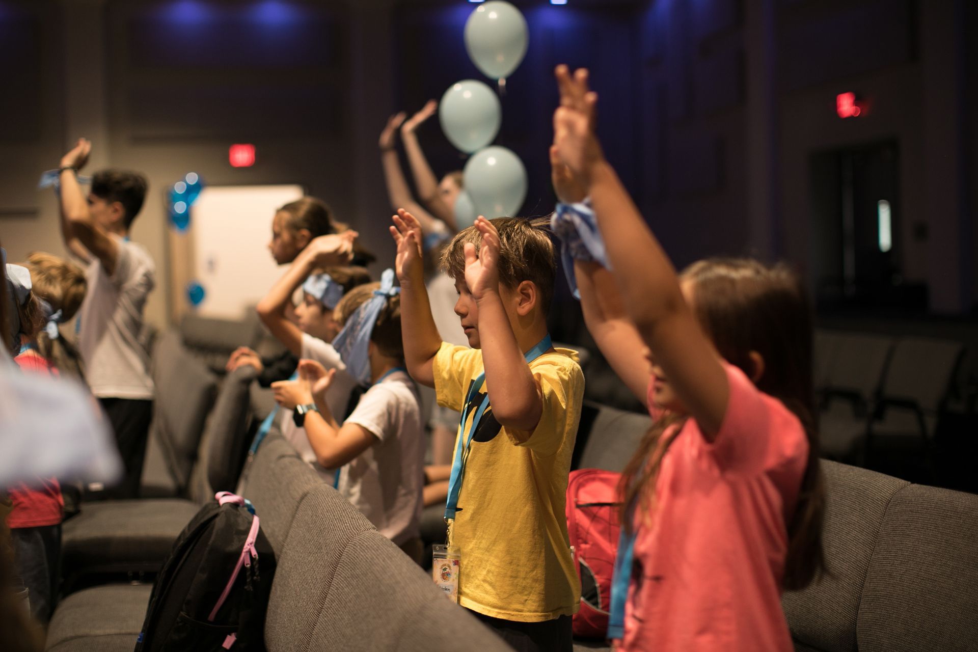 Group of people with hands raised in a dimly lit auditorium, with blue balloons floating above.