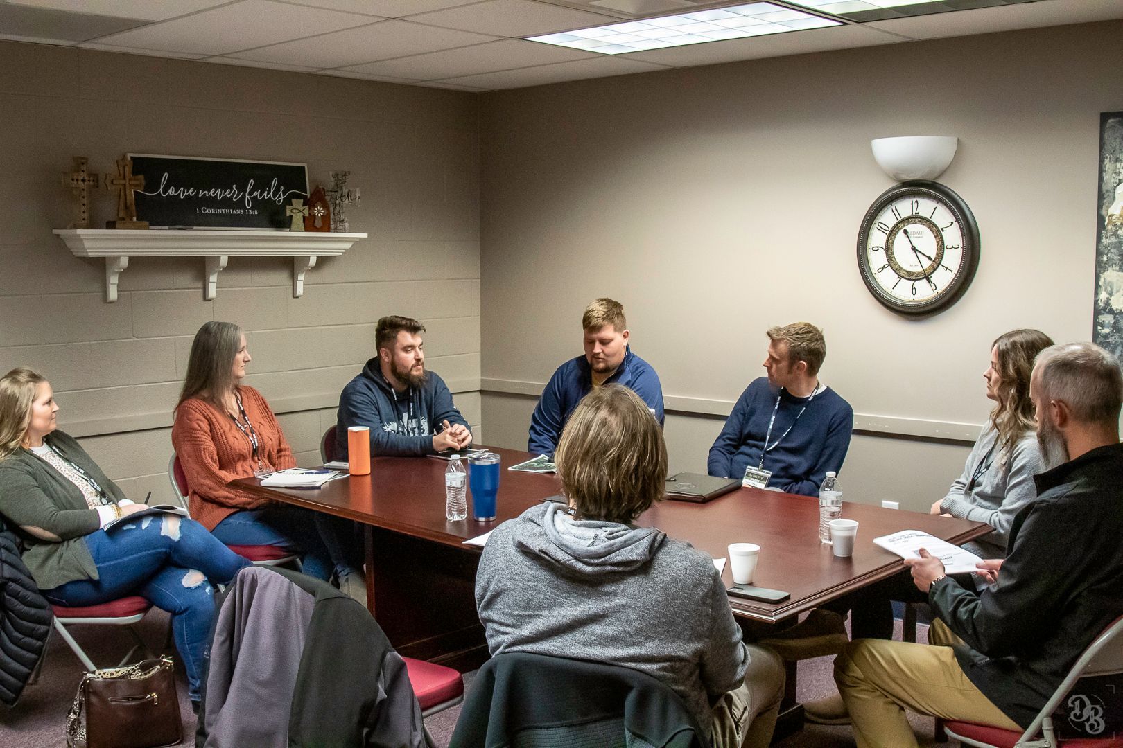 A group of eight people sitting around a long wooden conference table in a neutral-toned office meeting room.