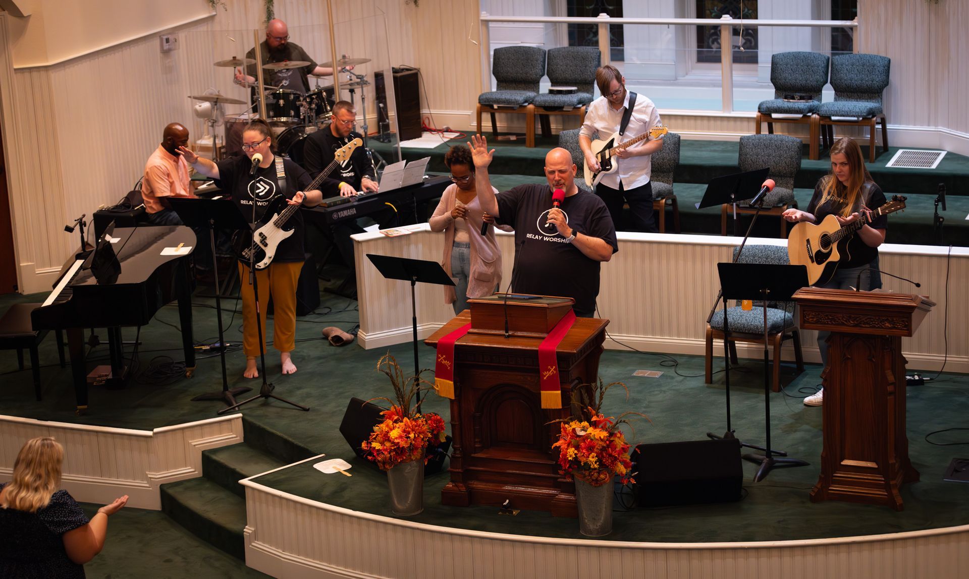 A band performs on a church stage, with members playing instruments and singing while a speaker stands at a podium.