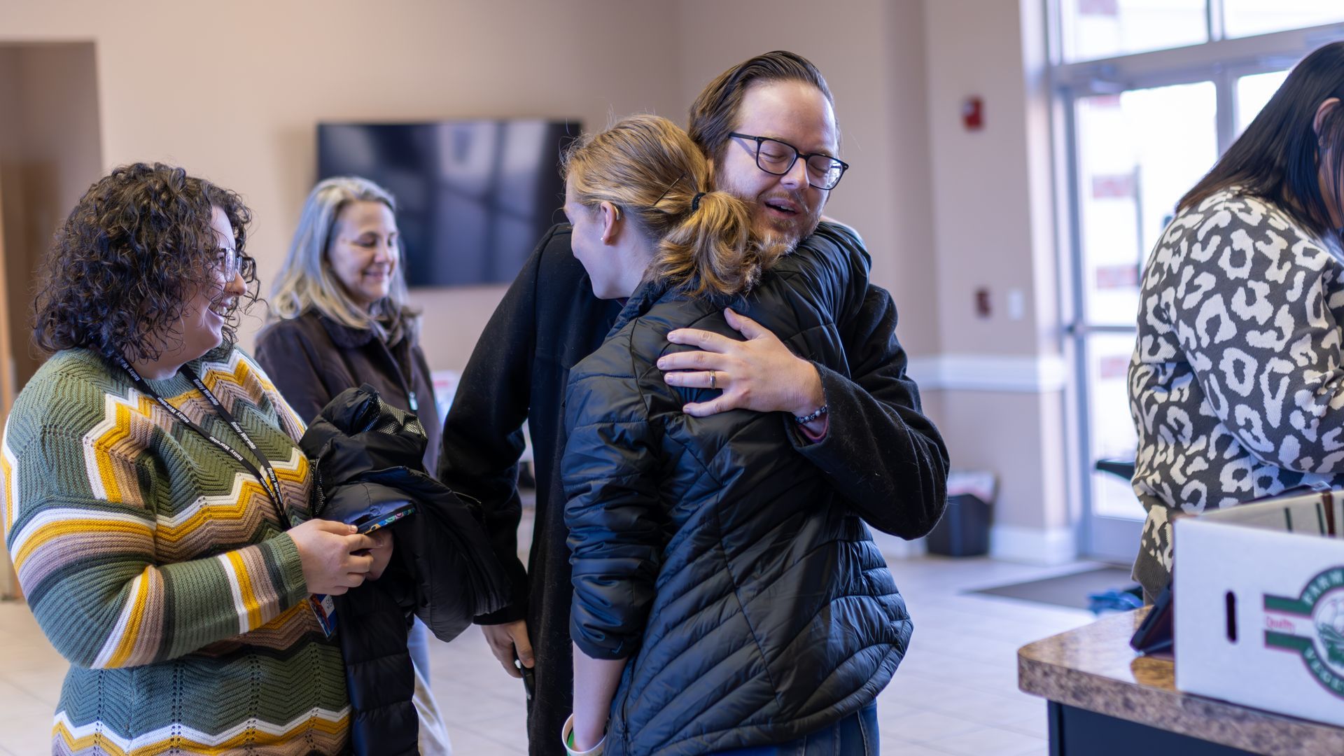A person in a black jacket hugs another person, while two others stand nearby in a bright, indoor setting.