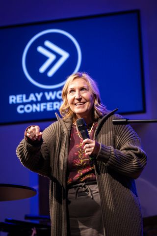 A speaker holds a microphone and gestures while presenting on stage at the Relay Women’s Conference.