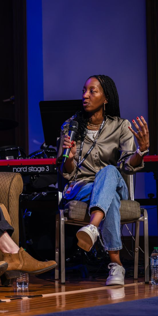 A person speaks into a microphone while seated on stage in front of a keyboard, gesturing with their left hand.