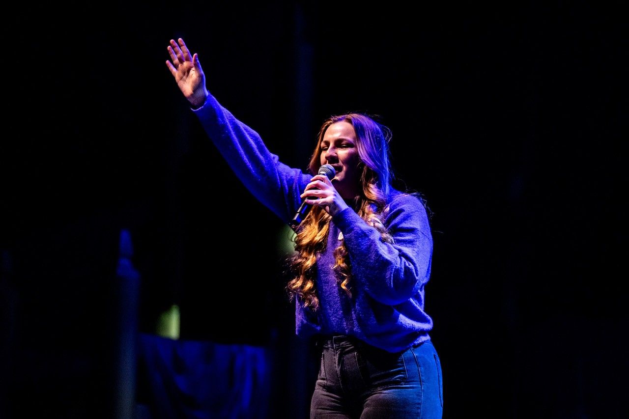 A person with long hair wearing a blue sweater, holding a microphone and waving to an audience on a dark stage.