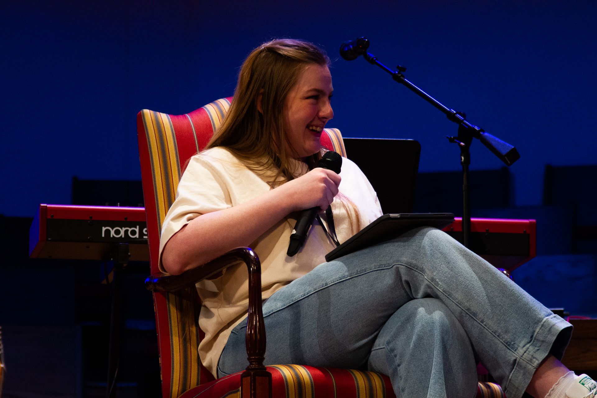 A person sits in a striped armchair on a stage, holding a microphone and a tablet, laughing against a blue background.