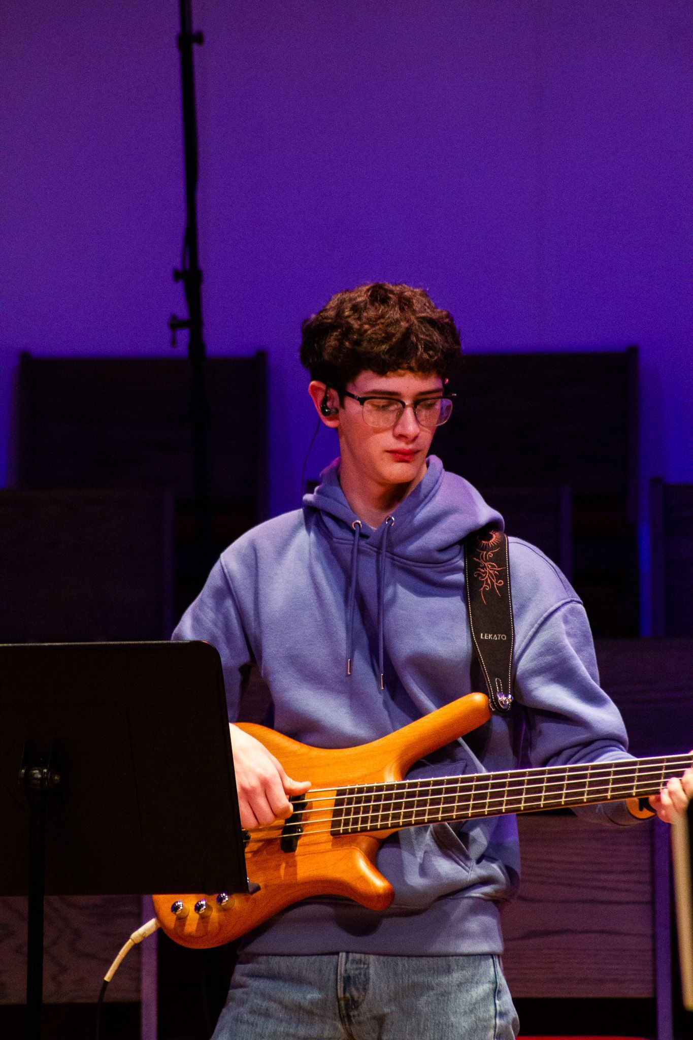 A person wearing glasses and a blue hoodie plays a tan bass guitar on a stage with a purple background.