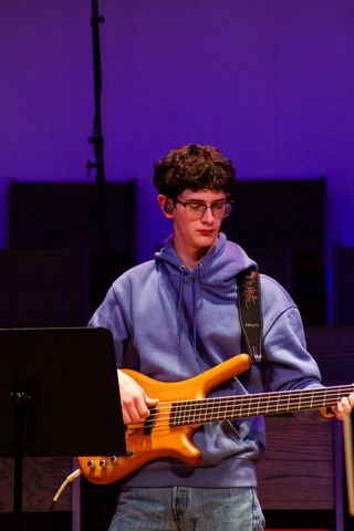 A person wearing glasses and a blue hoodie plays a tan bass guitar on a stage with a purple background.