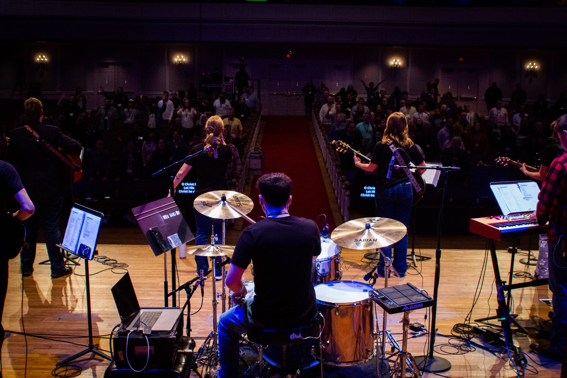 A band performs on a wooden stage for an audience seated in a darkened hall with a red-carpeted center aisle.