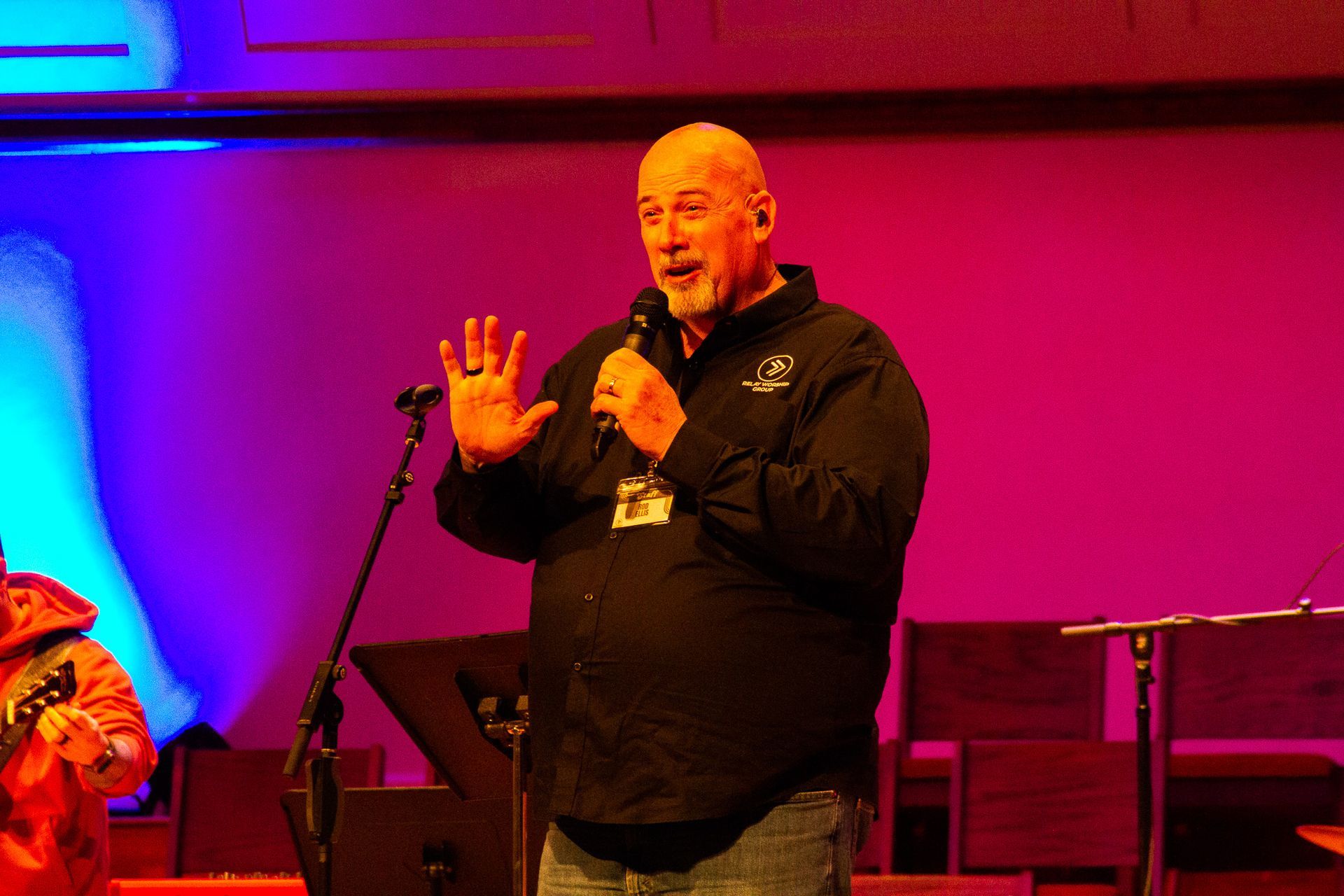 A person stands on a stage with red and blue lighting, speaking into a microphone while gesturing with one hand.