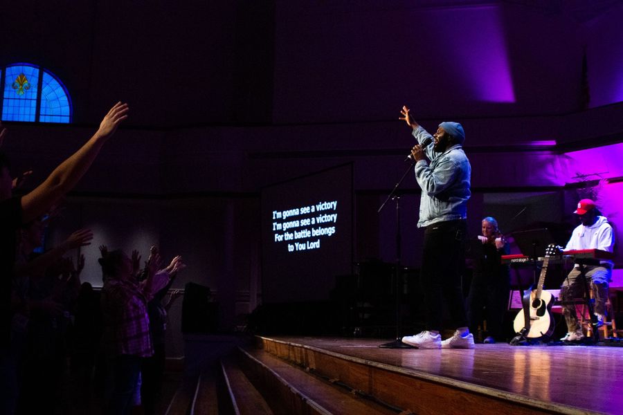 A singer performs on a stage in a dim, purple-lit church, with lyrics on a screen behind and audience members reaching up.