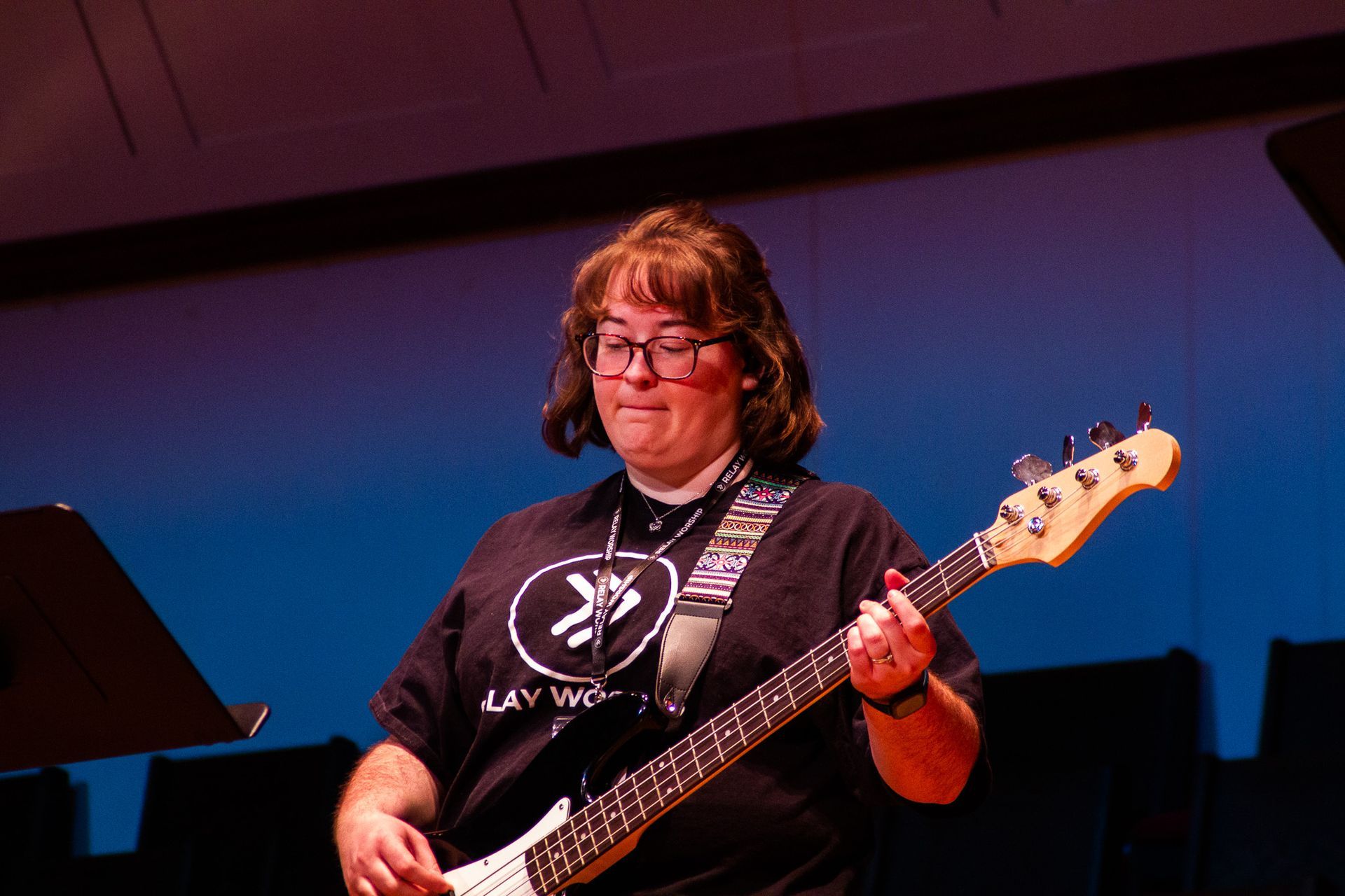 A person with shoulder-length brown hair and glasses plays a bass guitar on stage under blue and red lighting.