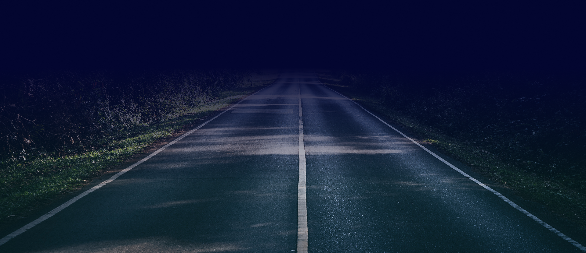 Dark, wet road at night with white dividing line. Grass and trees on the sides under a dark sky.