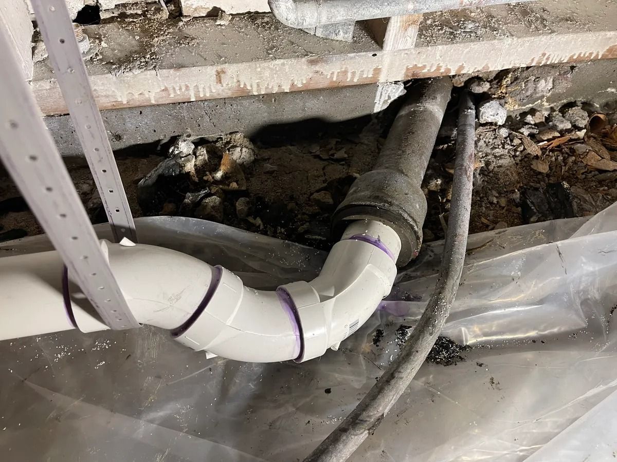 Exposed white PVC drain pipe with elbow joint under a damaged floor, surrounded by debris and plastic sheeting