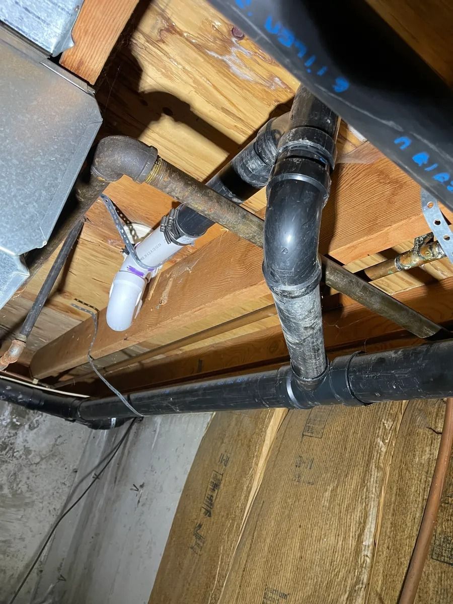 Under-sink plumbing with black drain pipes and a white PVC pipe beneath a wooden floor.