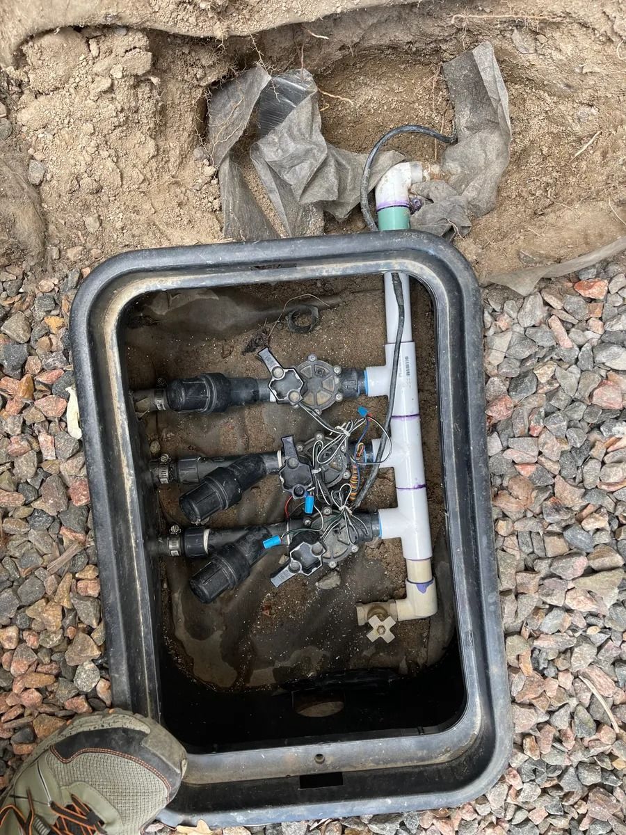 Open water meter box with exposed pipes and valves in gravel and dirt ground