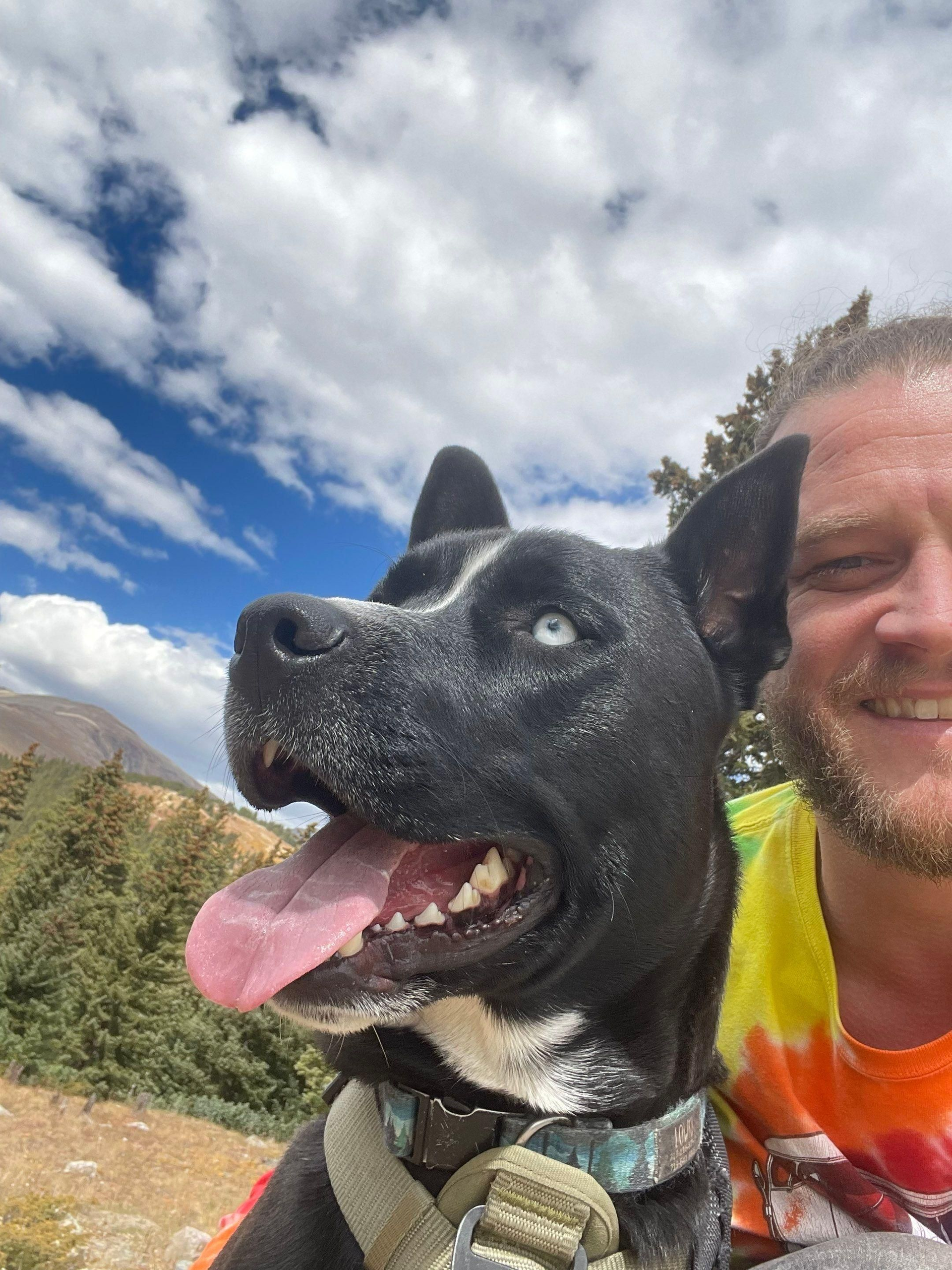 Black dog with tongue out beside a smiling person in a tie-dye shirt under a cloudy sky