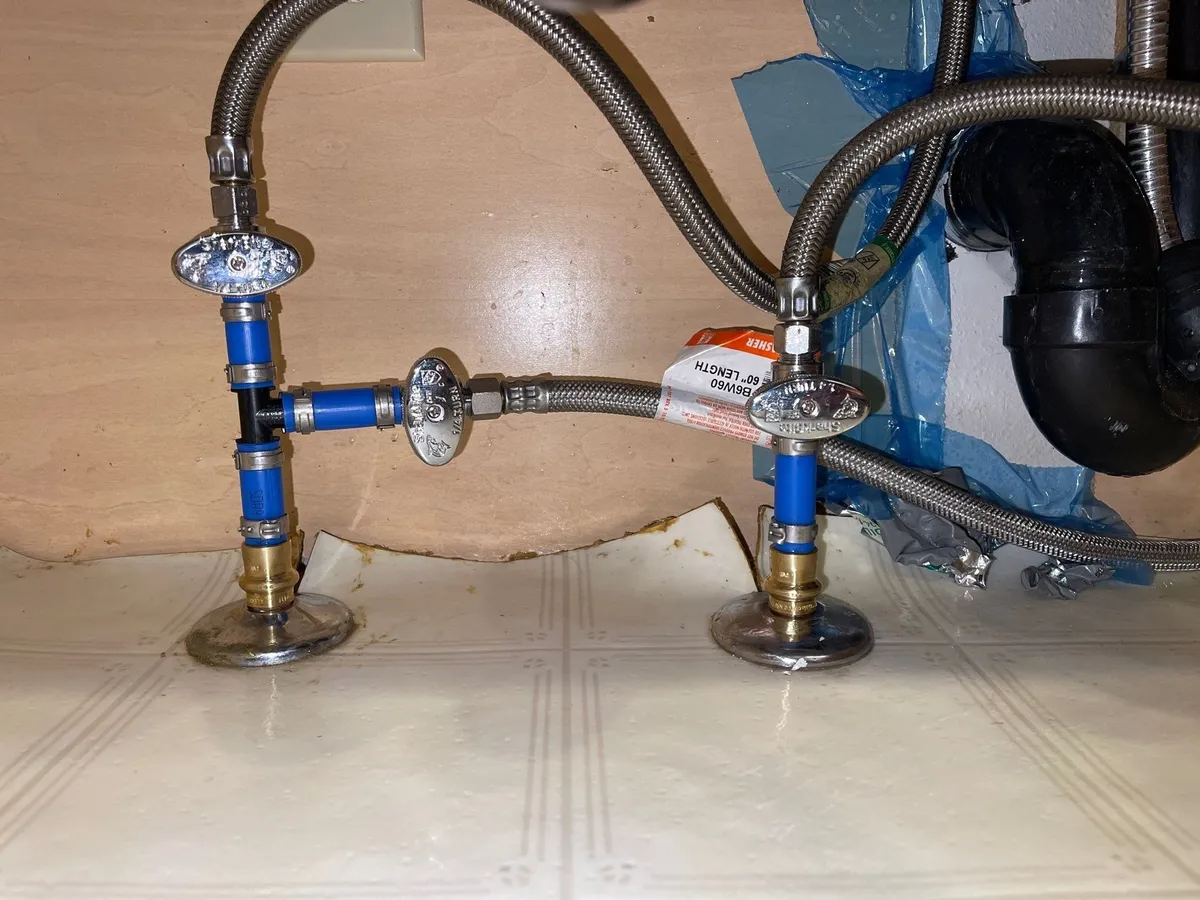 Under-sink plumbing with blue valves, braided hoses, and a leak-damaged cabinet floor