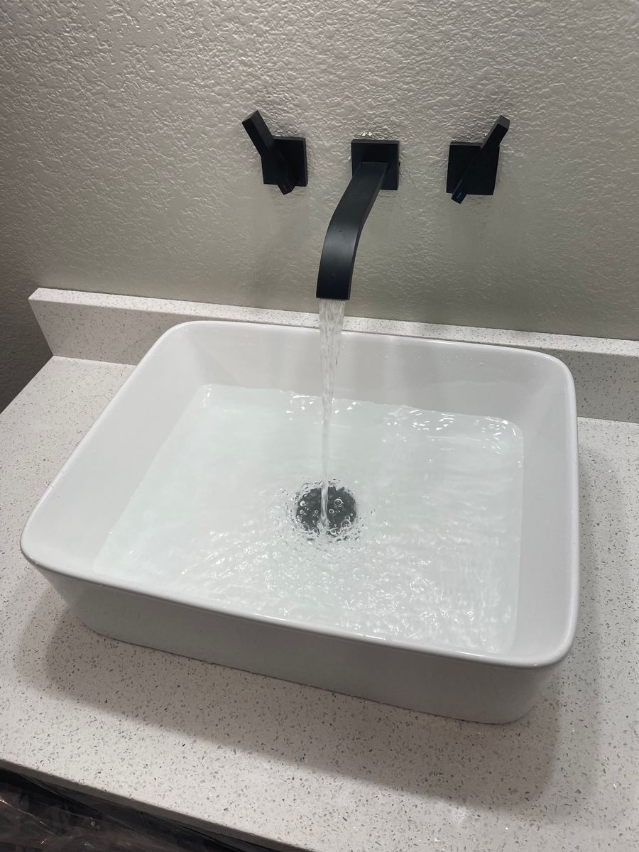 Water flowing from a black wall faucet into a white rectangular sink on a countertop.