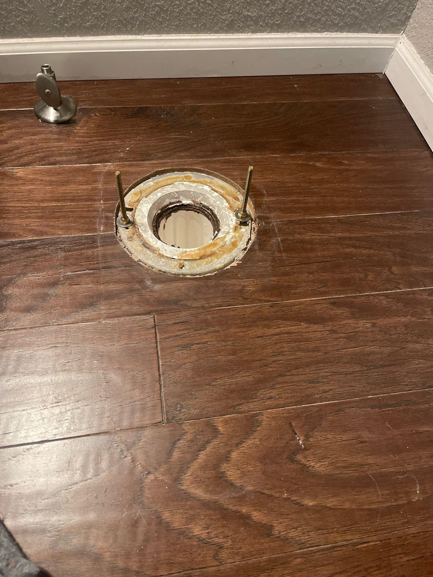 Floor cutout with exposed pipe hole in dark wood flooring near the wall