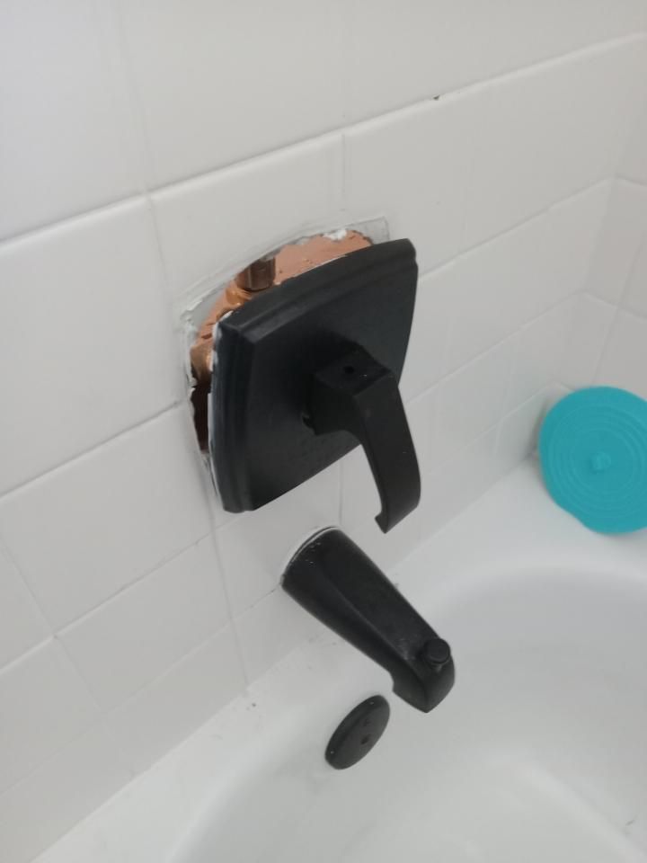 Broken black bathtub faucet mounted on white tile, with exposed copper pipe and detached handle.