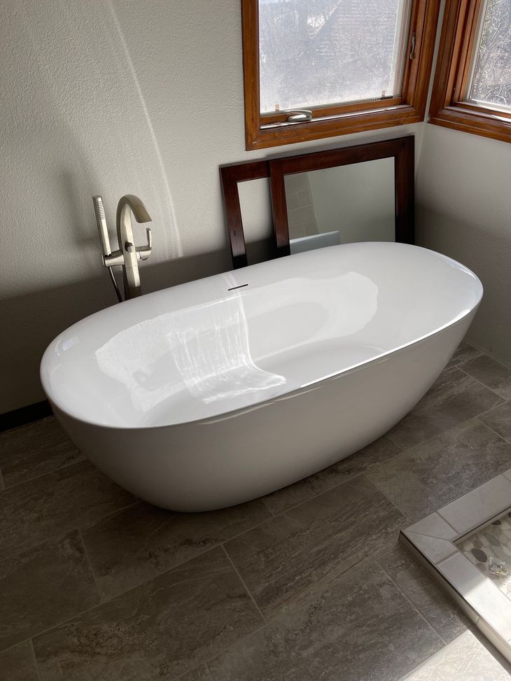 Modern white freestanding bathtub beside a window in a tiled bathroom