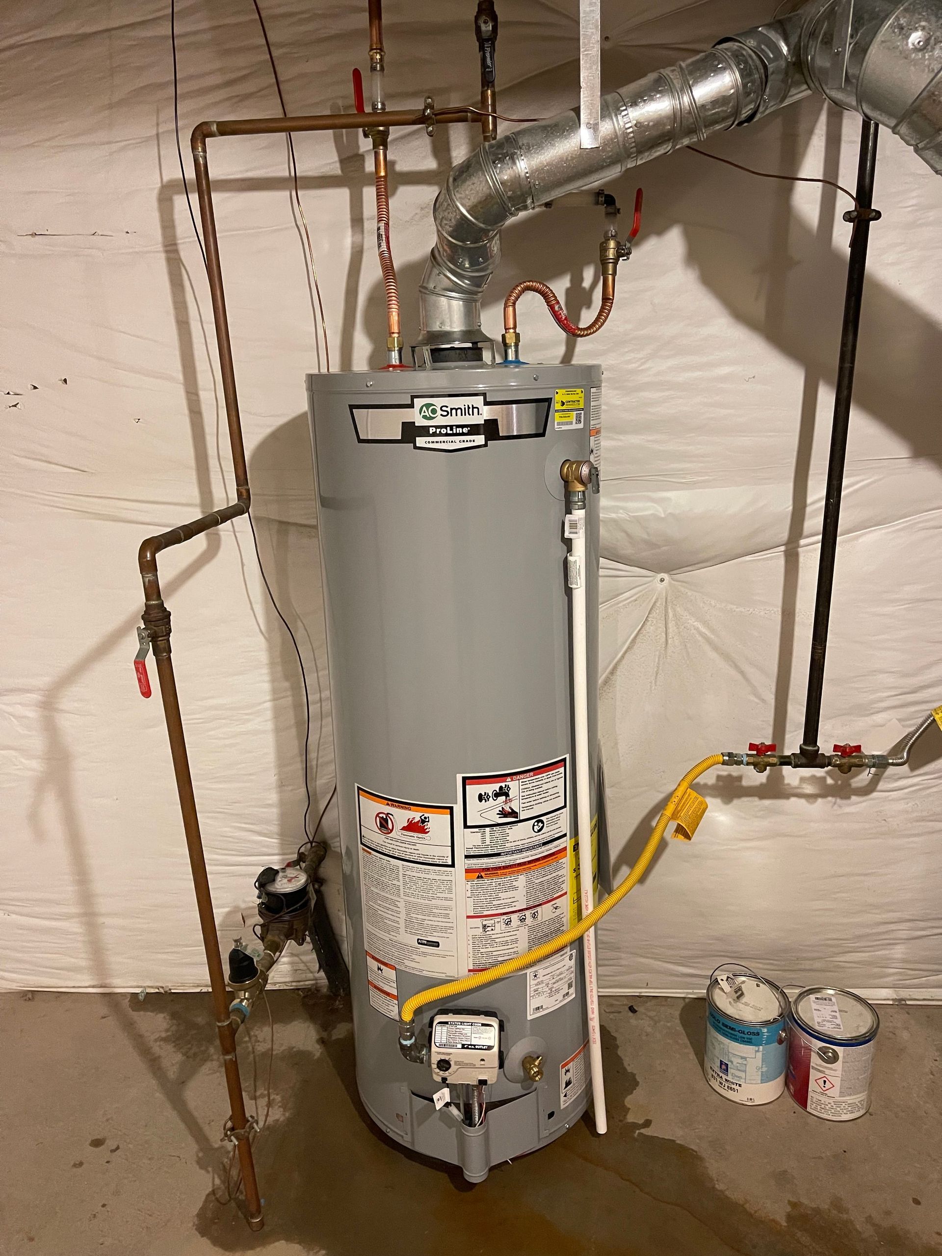 Water heater in a basement with exposed pipes and a gas line, next to paint cans.