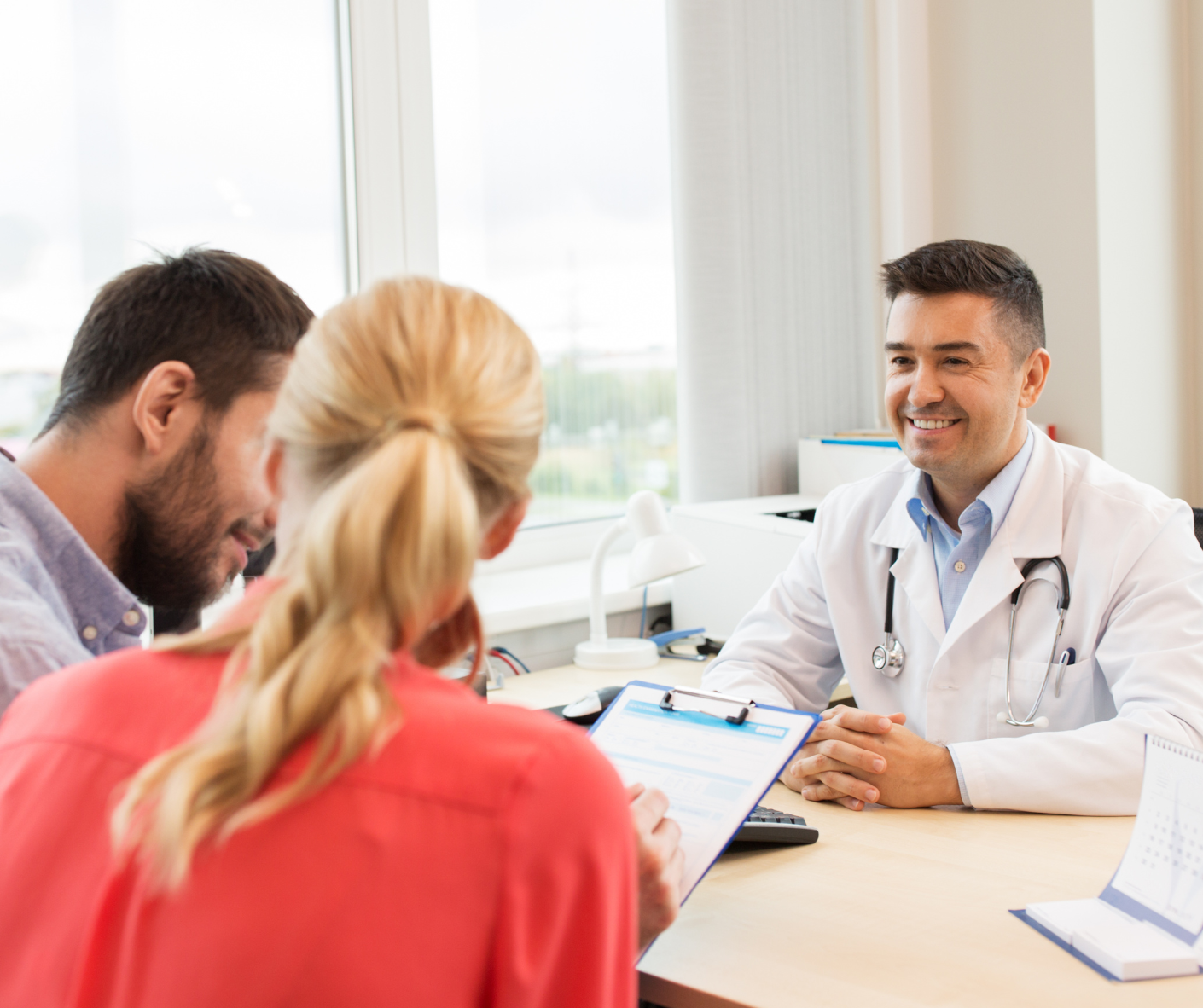 Doctor smiling at a couple seated at a desk; doctor's office setting with a clipboard.