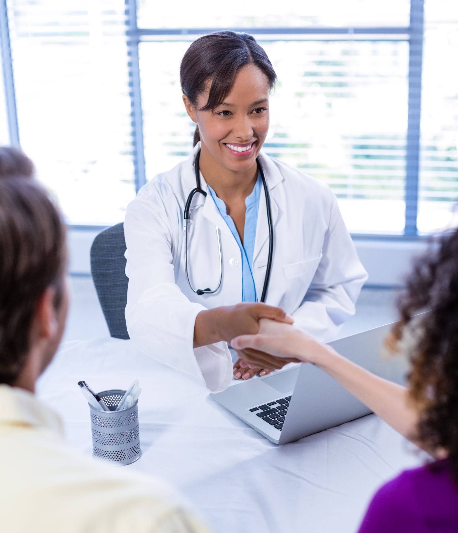 A doctor is shaking hands with a patient while sitting at a desk with a laptop.