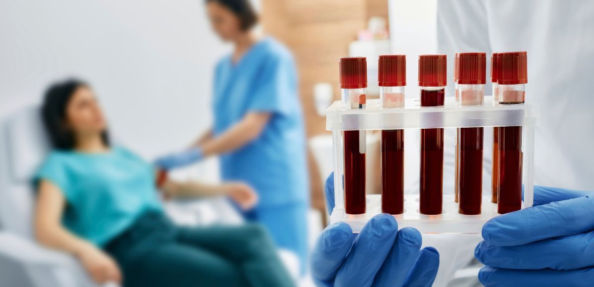 Gloved hands holding test tubes of blood samples in a clinical setting with a patient in the background