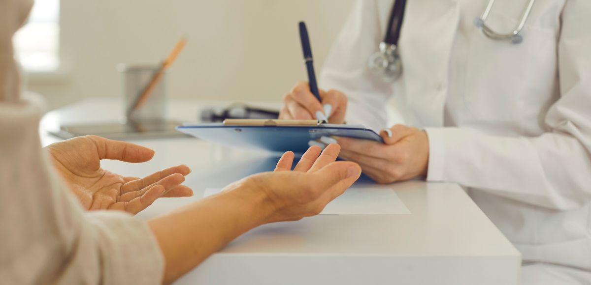 Doctor writing on a clipboard while speaking with a patient’s outstretched hands in a bright clinic