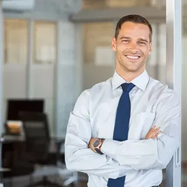 A man in a white shirt and blue tie is standing in an office with his arms crossed and smiling.