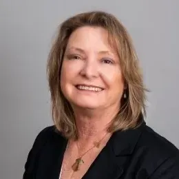 Woman with shoulder-length blonde hair, wearing a black blazer, smiles. Gray background.