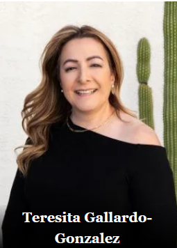 Woman with long wavy brown hair, smiling, wearing black top, posing outdoors with cactus.