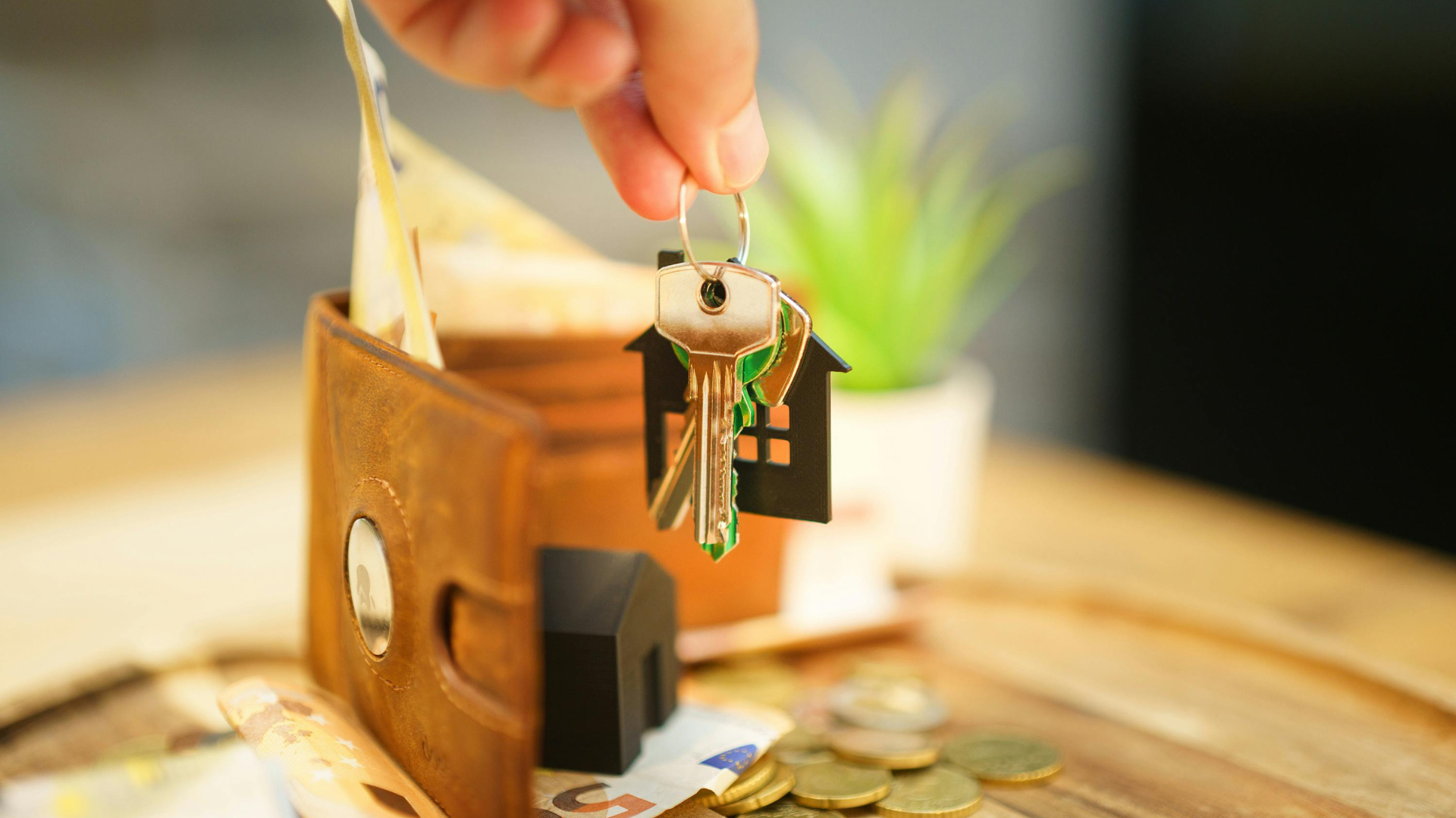 Hand holding keys with a house-shaped charm above a wallet with cash, coins, and a plant.