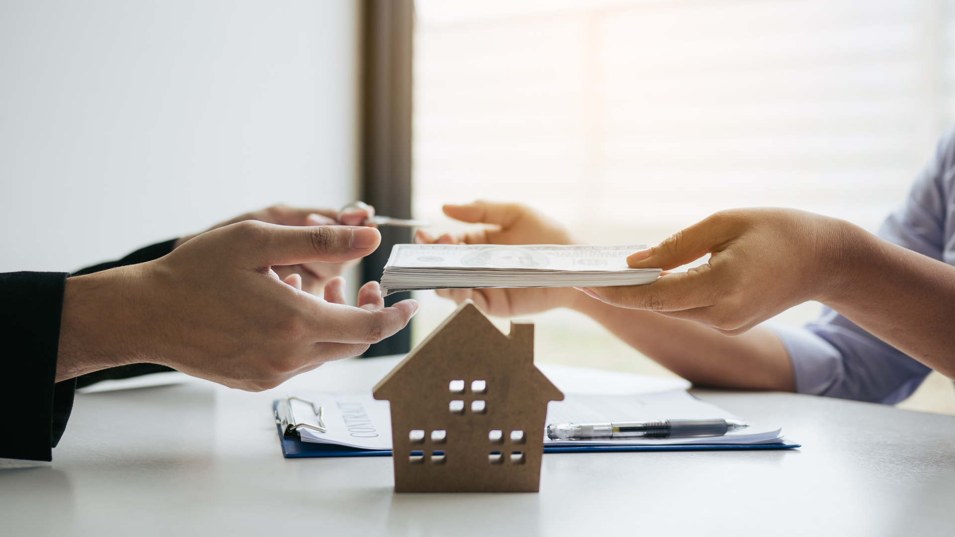 Hands exchanging money over a small house model on a table with documents and a pen.
